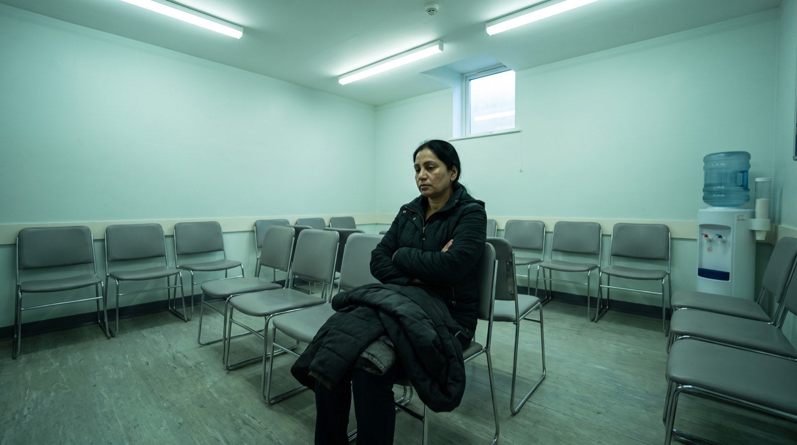 A woman waiting alone in a row of chairs in a clinic waiting area under fluorescent light
