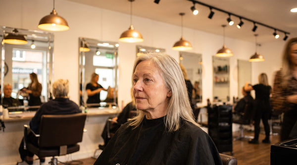 A woman in her sixties waiting in a hairdresser's chair, no makeup, sparse pale lashes