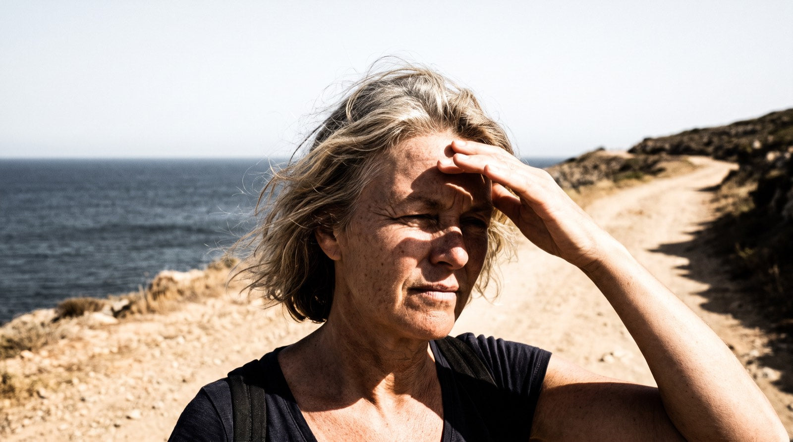 A woman in her late fifties squinting into hard midday sun on a coastal footpath