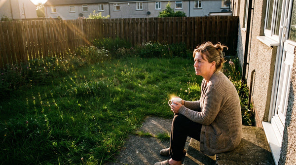 A woman sitting on the back step of a house with a mug of tea in low evening sun