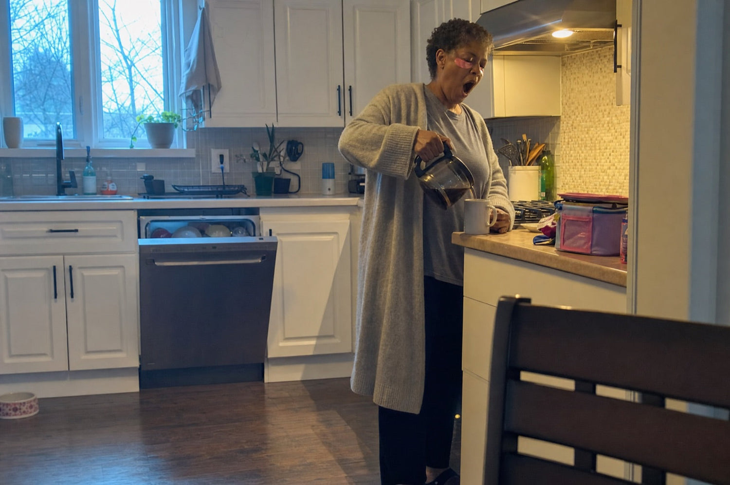 Woman in her fifties pouring coffee in a kitchen at dawn with pink hydrogel patches under her eyes